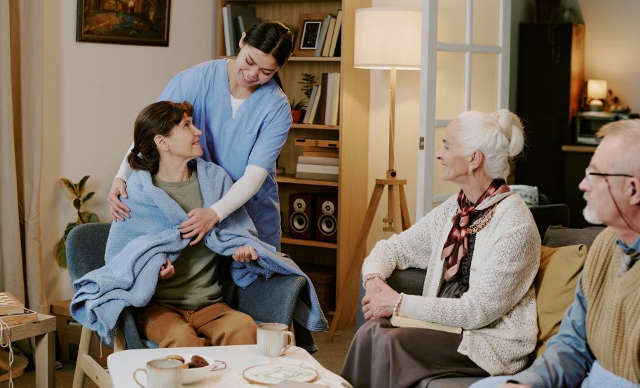 A smiling young female nurse in blue scrubs gently wraps a cozy blue blanket around a seated senior woman in a living room, while an elderly couple watches warmly from a nearby sofa.