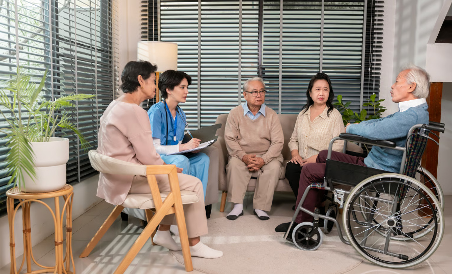 An Asian female nurse in blue scrubs sits with a clipboard, listening to an elderly Asian man in a wheelchair during a group health discussion with three other senior residents in a modern, sunlit living room.