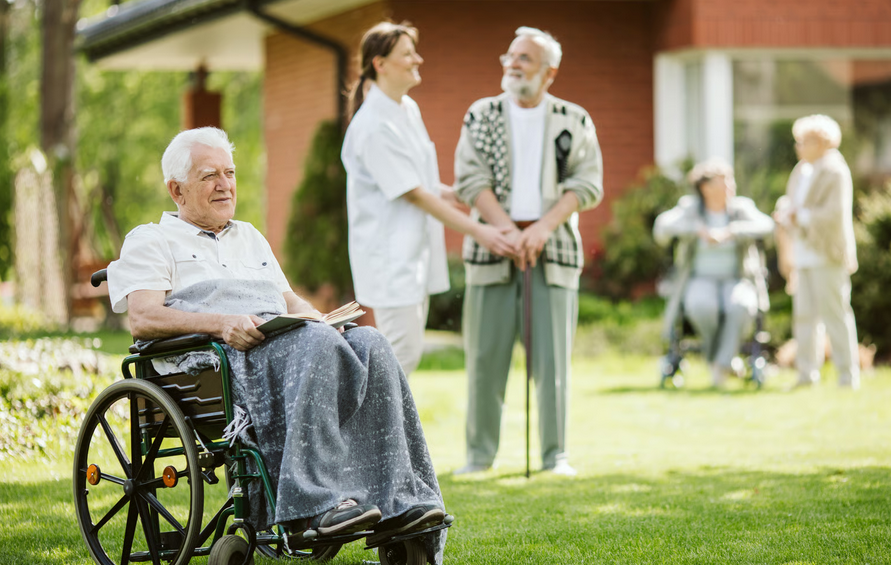 An elderly man in a wheelchair holds a book on a sunny lawn, smiling peacefully while a caregiver and other seniors interact in the background. This highlights how early hospice enrollment focuses on dignity and comfort, allowing patients more meaningful time outdoors and at home.