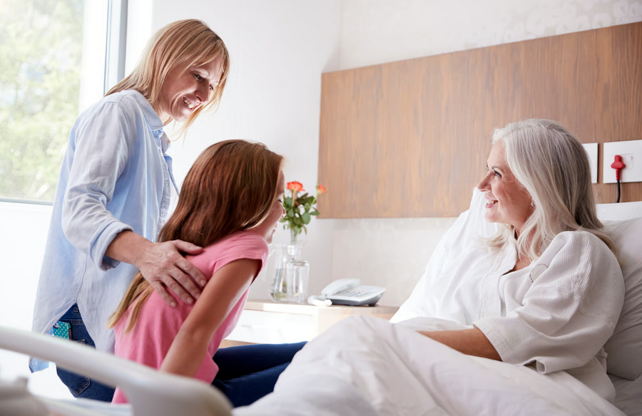 A mother and daughter smile warmly during a visit with an elderly woman resting comfortably in a hospital bed at home. This scene exemplifies what families can expect in the first 48 hours of hospice care, as localized clinical support allows families to step away from crisis management and focus entirely on being present with the people they love.
