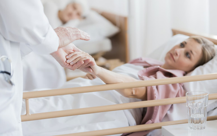 A medical professional in a white coat holding the hand of a female patient resting in an inpatient facility bed.