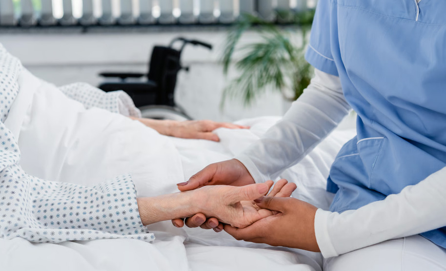 A close-up of a hospice caregiver in blue scrubs gently holding the hand of a patient resting in a hospital bed, with a wheelchair softly blurred in the background, illustrating the hands-on clinical comfort and support provided under the Medicare six-month prognosis guidelines.