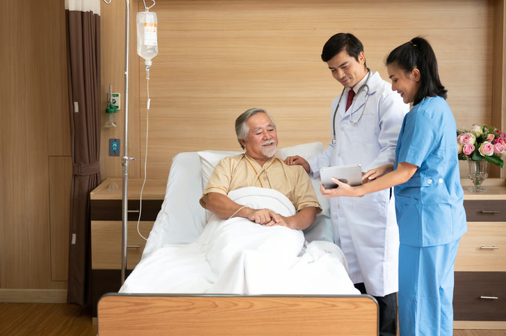 A male doctor and female nurse in blue scrubs discussing a digital tablet with a senior male patient resting in a hospital bed.