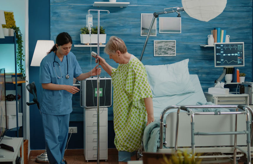 Hospice nurse helping a senior female patient walk using an IV pole alongside a medical bed and walker at home.