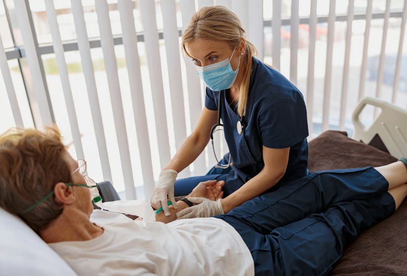 A nurse wearing a surgical mask and gloves attending to an elderly patient in a medical bed.