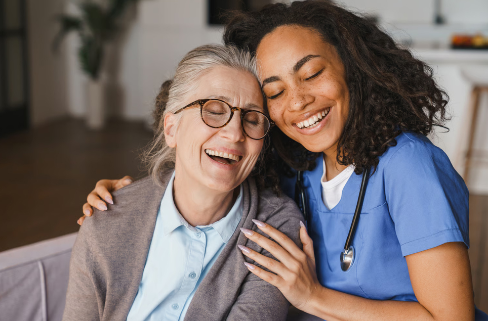 A compassionate healthcare worker in blue scrubs embracing and laughing with an elderly female patient receiving palliative care.